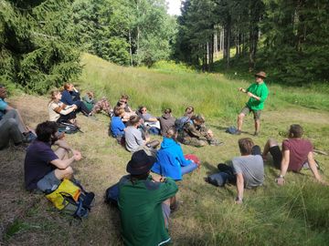 Eine Menschengruppe sitzt auf einer Bergwiese und hört dem Freiwilligen Stefan Escher zu.
